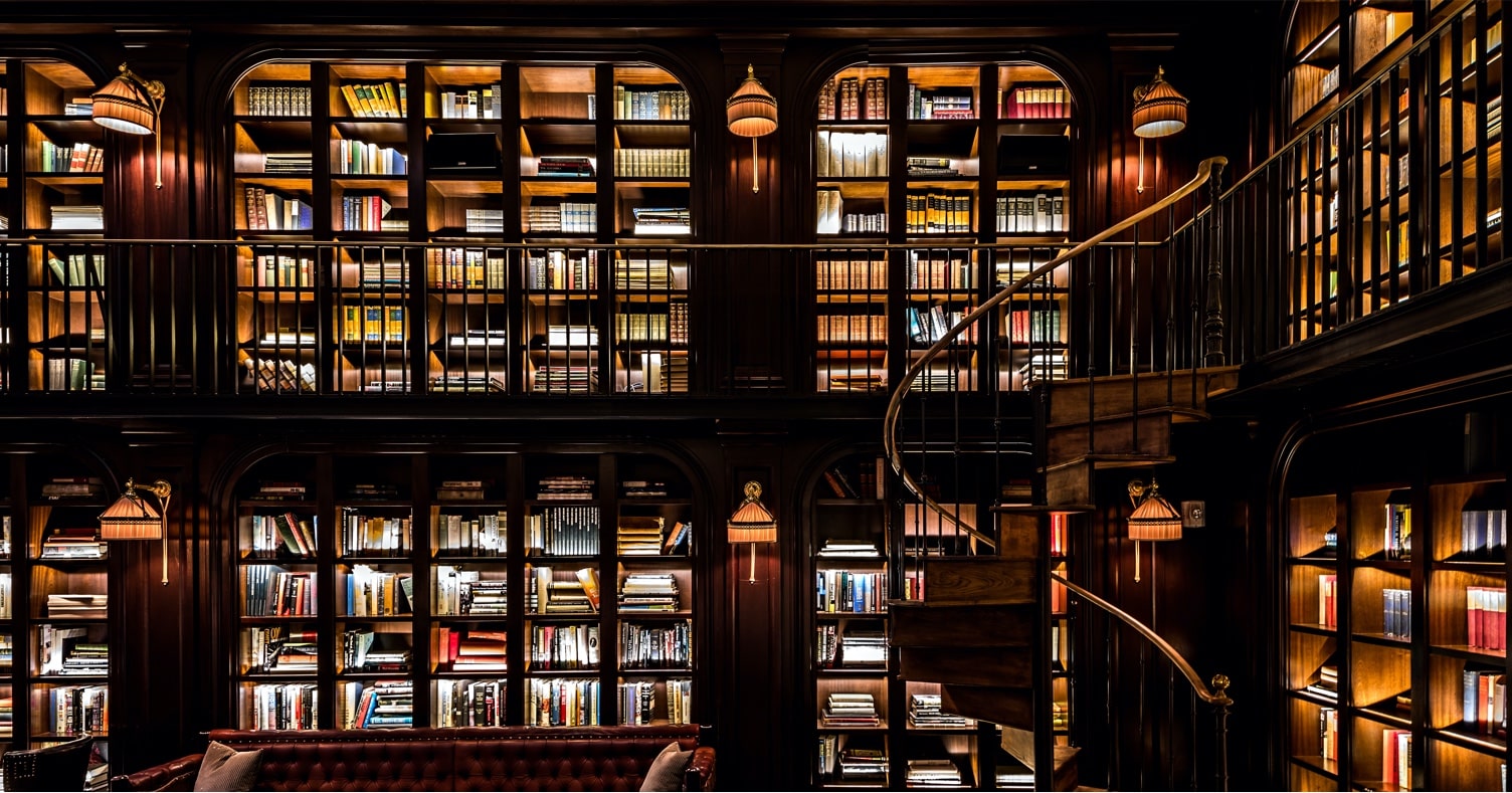 Lowlit room in the Library of the NoMad hotel, showcasing the wall-to-wall bookcases and spiral staircase.