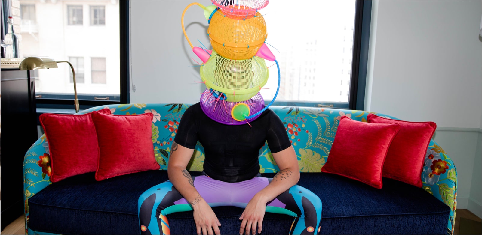 Gentleman sitting on a couch wearing colorful pants with a sculpture on his head