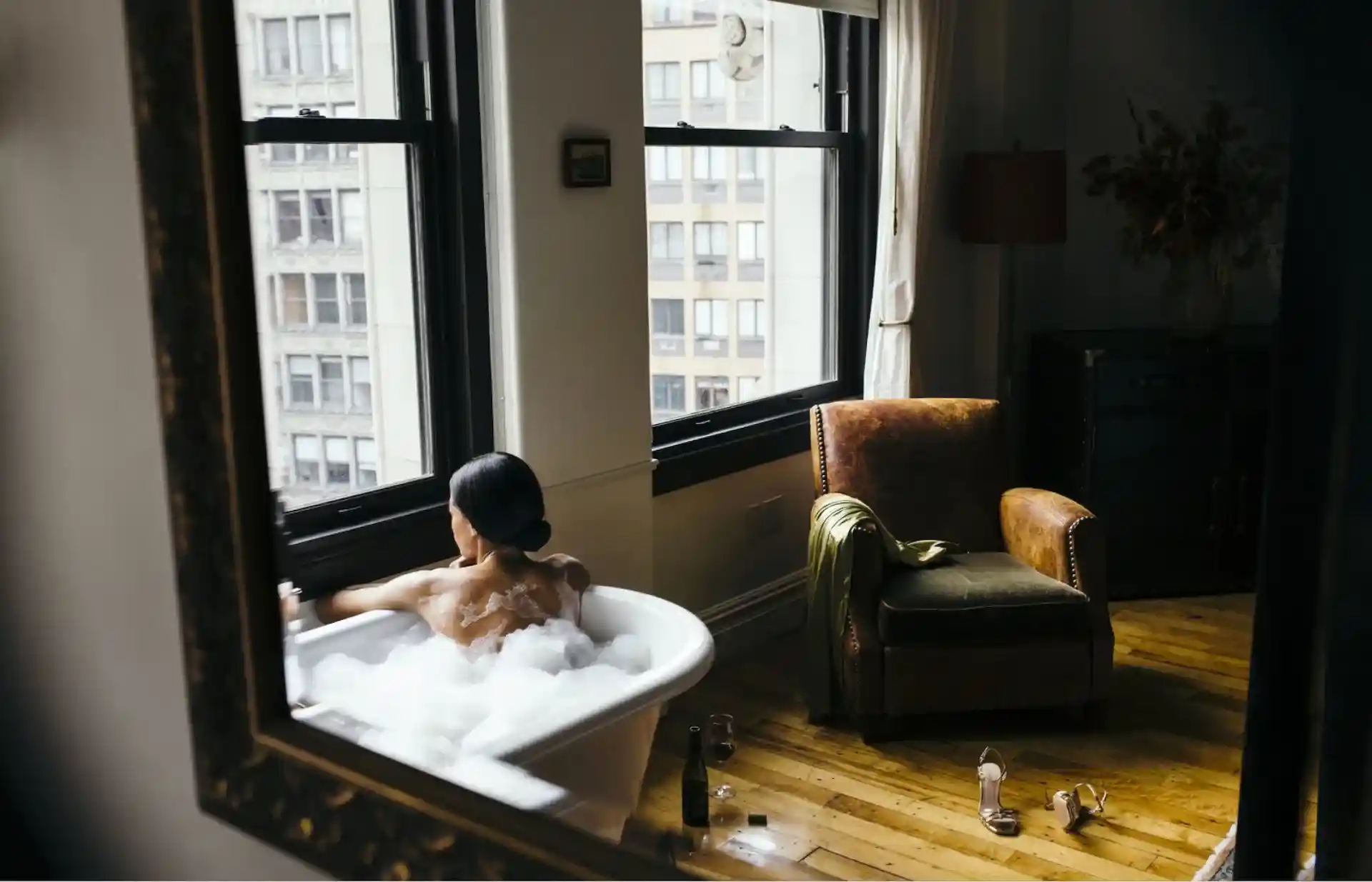 Mirror reflects a woman with her back facing the viewer in a bubble filled bathtub, looking out a window overlooking the city.